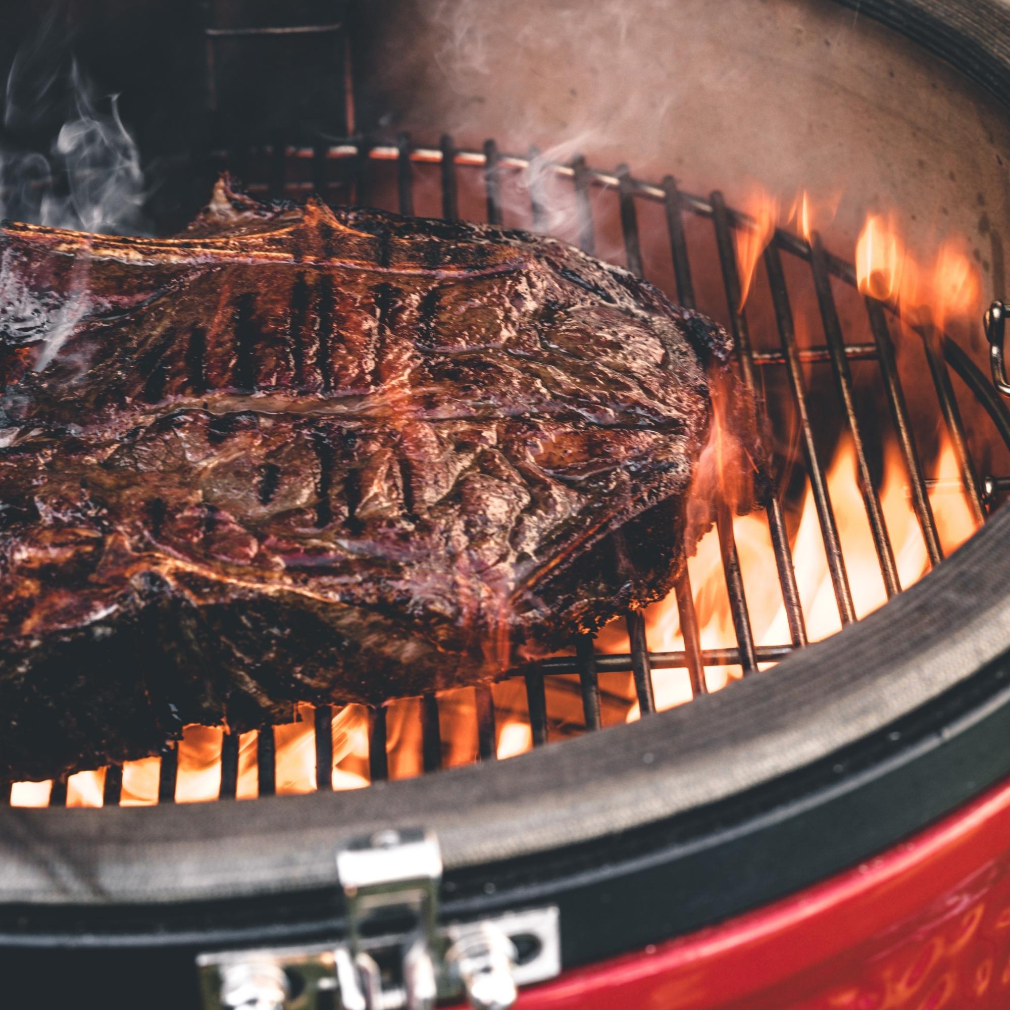 Großes, gebratenes Steak liegt auf dem Grillrost eines Holzkohlegrills mit sichtbarer Flamme