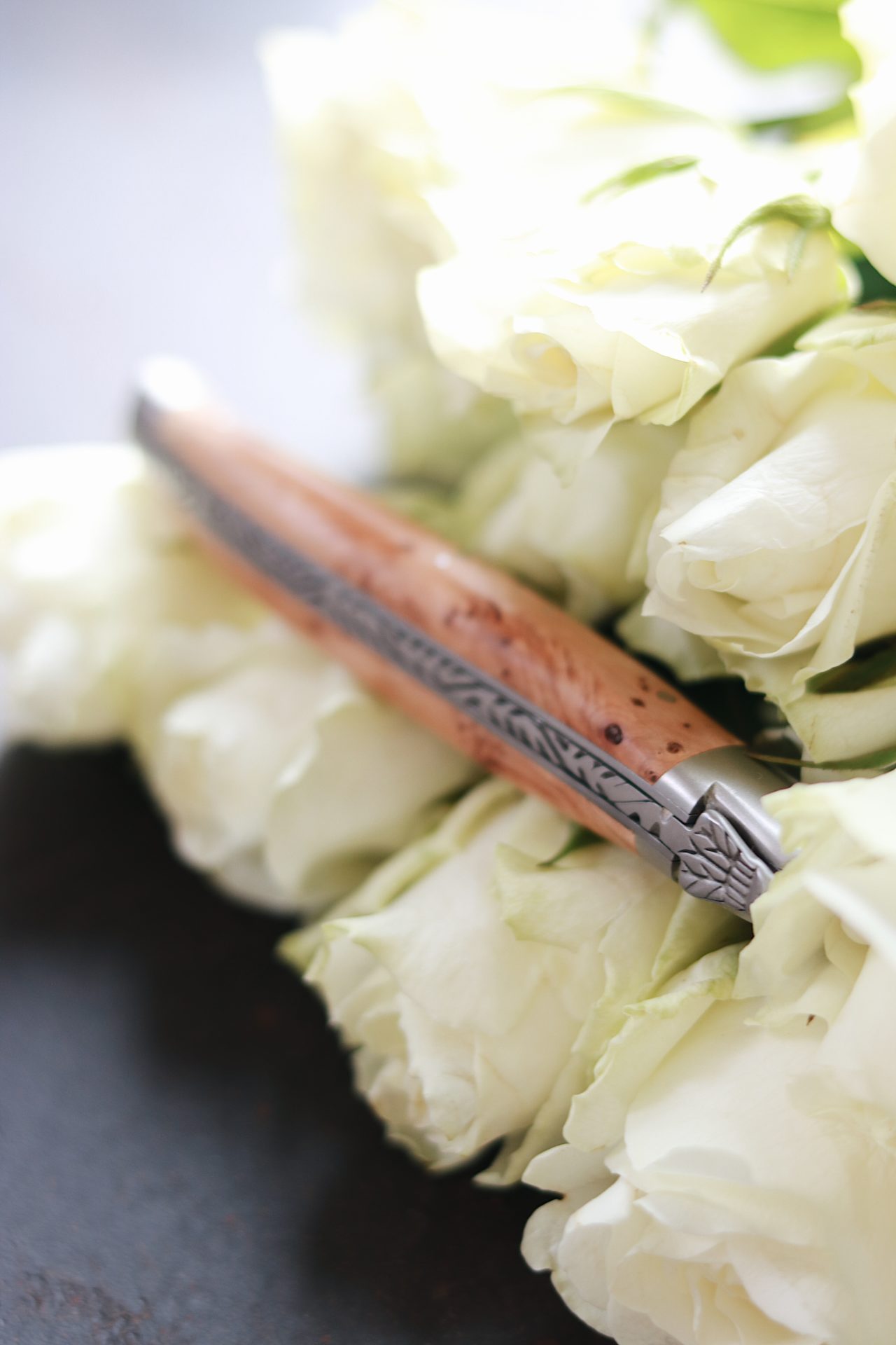 Detailaufnahme eines Klappmessers mit verziertem Metall und Holzgriff, liegend auf mehreren weißen Rosenblüten