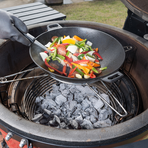 Eine Person kocht buntes Gemüse und Wurst in einem Wok auf einem Keramik-Grill über glühender Holzkohle