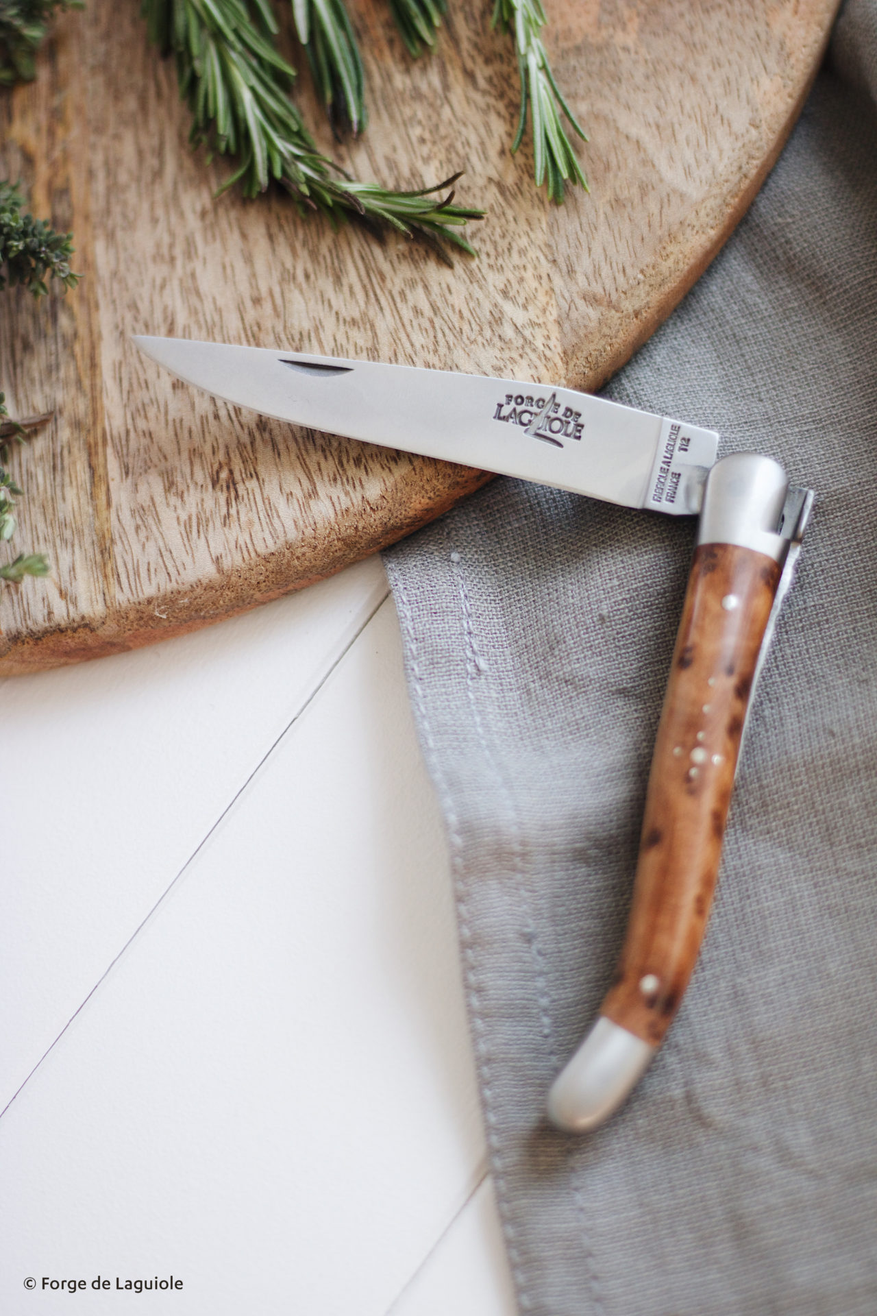 Elegantes Laguiole Klappmesser mit Holzgriff, liegend auf grauer Serviette und Holzbrett, mit frischem Rosmarin und Thymian