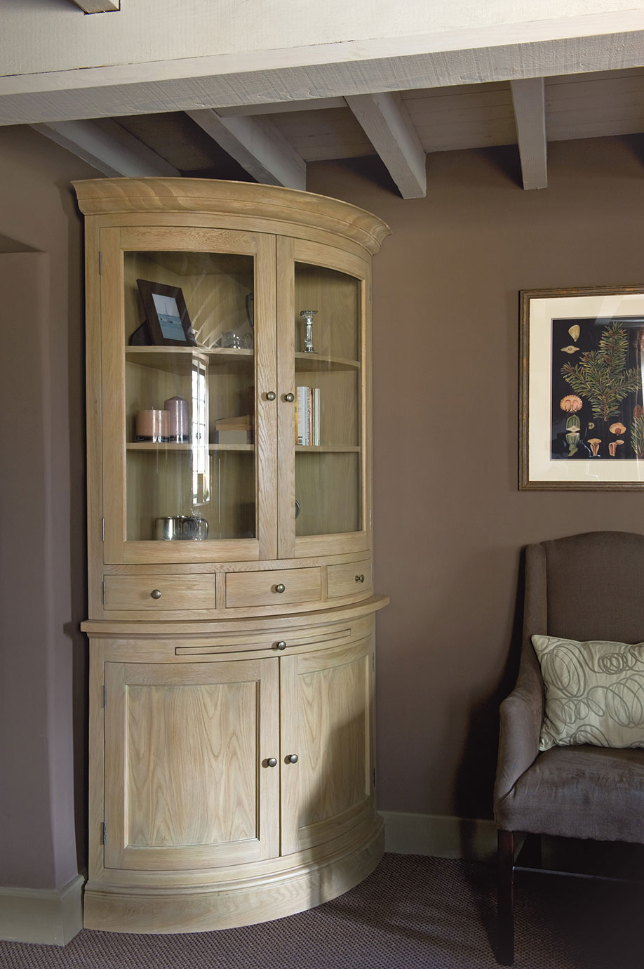 Eckvitrine aus Massivholz mit Glastüren, mehreren Regalböden und Schubladen, im Raum zwischen Sessel und Wand platziert