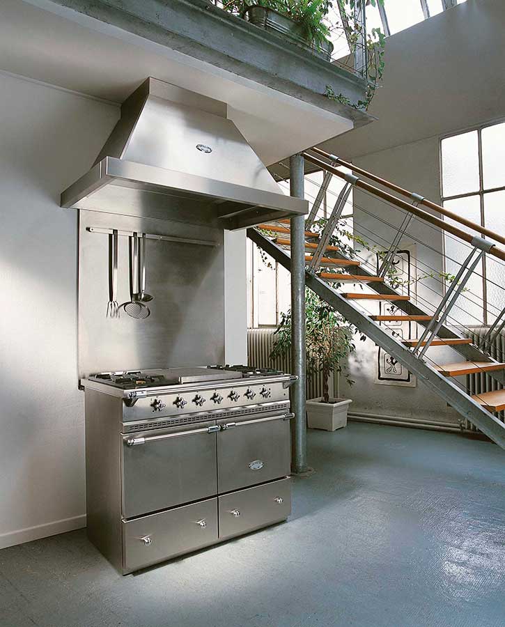 Moderner Edelstahlherd mit Kochutensilien und großer Abzugshaube in loftartiger, heller Küche mit offener Treppe und Pflanzen