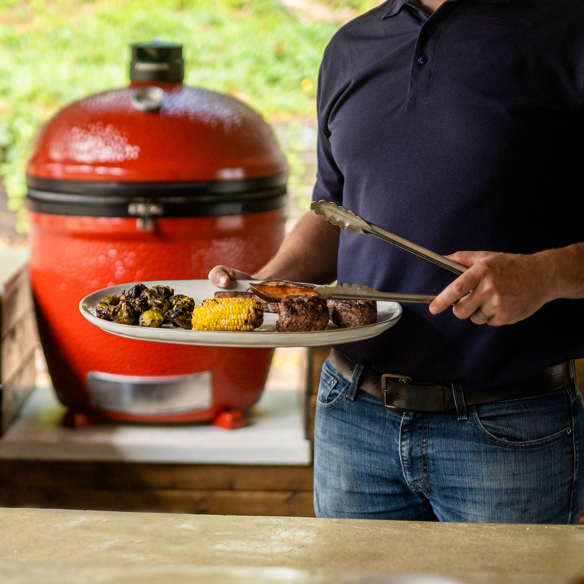 Person hält eine Grillplatte mit Fleisch, Maiskolben und gegrilltem Gemüse vor einem roten Keramikgrill im Freien