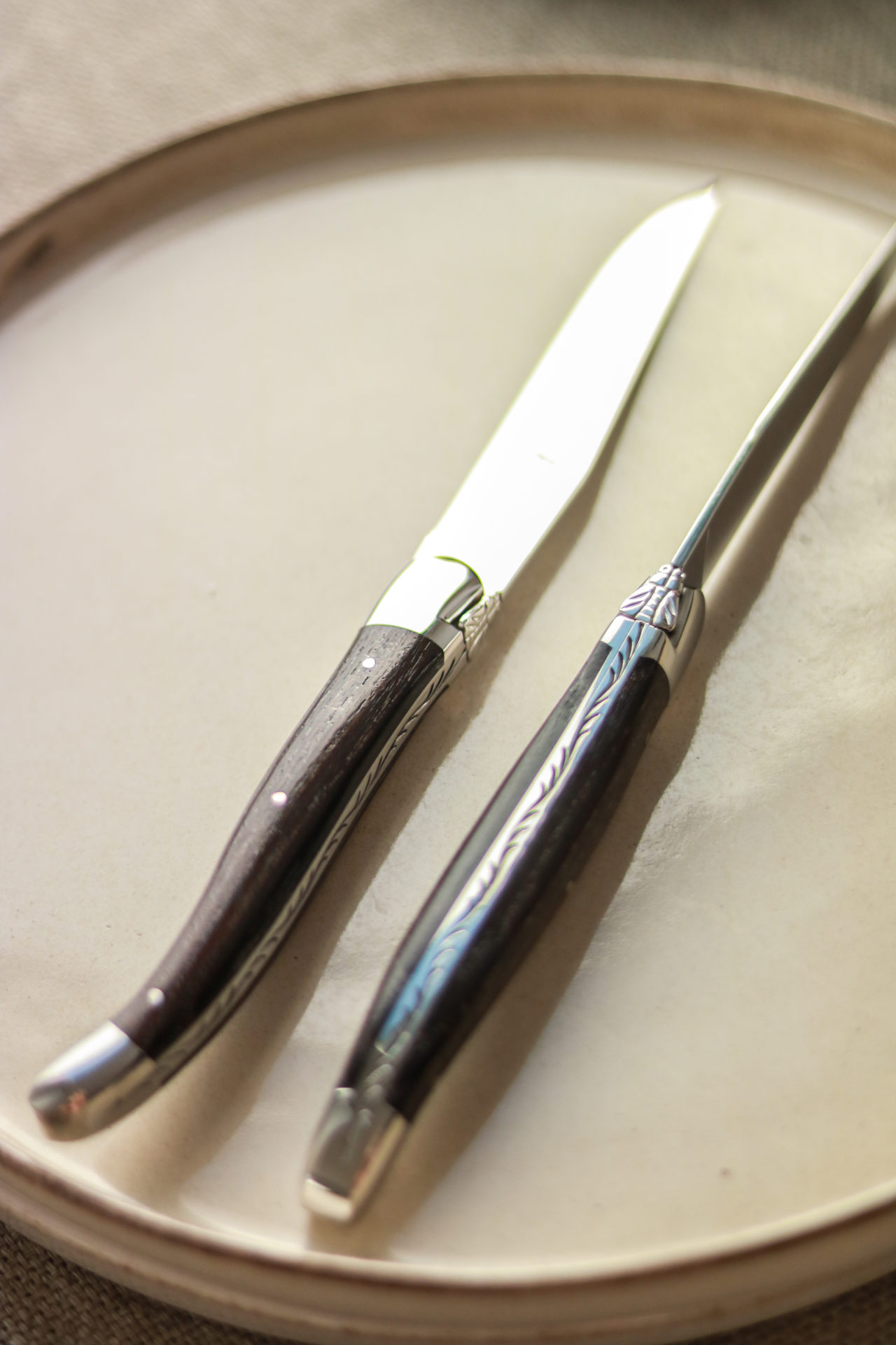 Zwei hochwertige Steakmesser mit dunklem Holzgriff und verziertem Metall auf einem hellen Teller