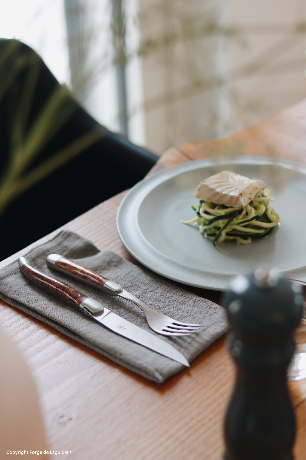 Detailansicht eines gedeckten Tisches mit einem hochwertigen Messer und einer Gabel mit Holzgriff, serviert auf einer grauen Stoffserviette, daneben ein Teller mit Fischfilet auf Gemüsenudeln