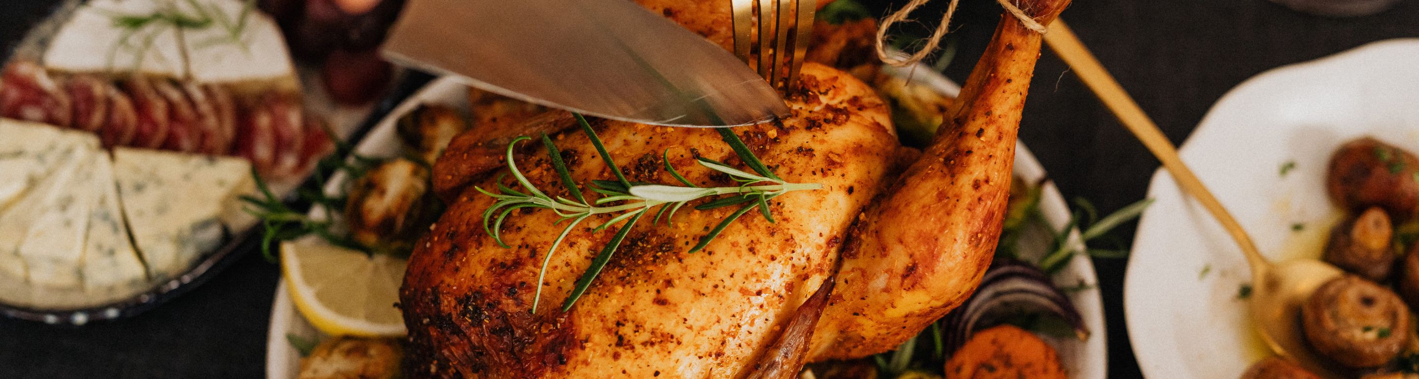 Knusprig goldbraunes Brathähnchen mit Rosmarin wird aufgeschnitten; serviert mit Zitronenscheiben, Ofengemüse und kleinen Kartoffeln, daneben Käse- und Wurstplatte