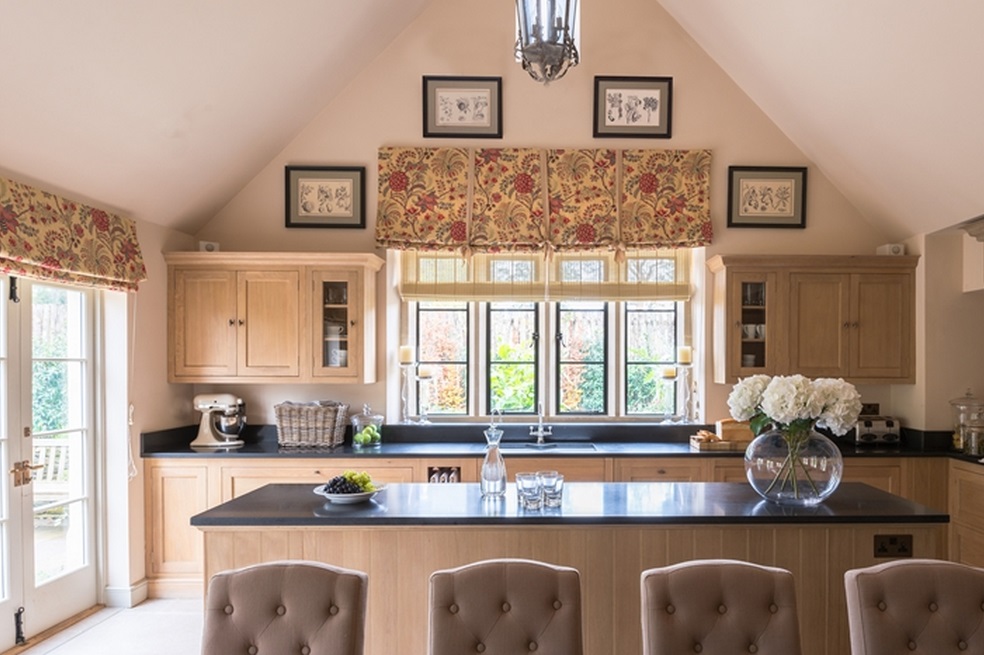 Gemütliche Küche im Landhausstil mit Holzschränken, floral gemusterten Rollos, Kücheninsel und Blumendekoration in einem großen, hellen Raum