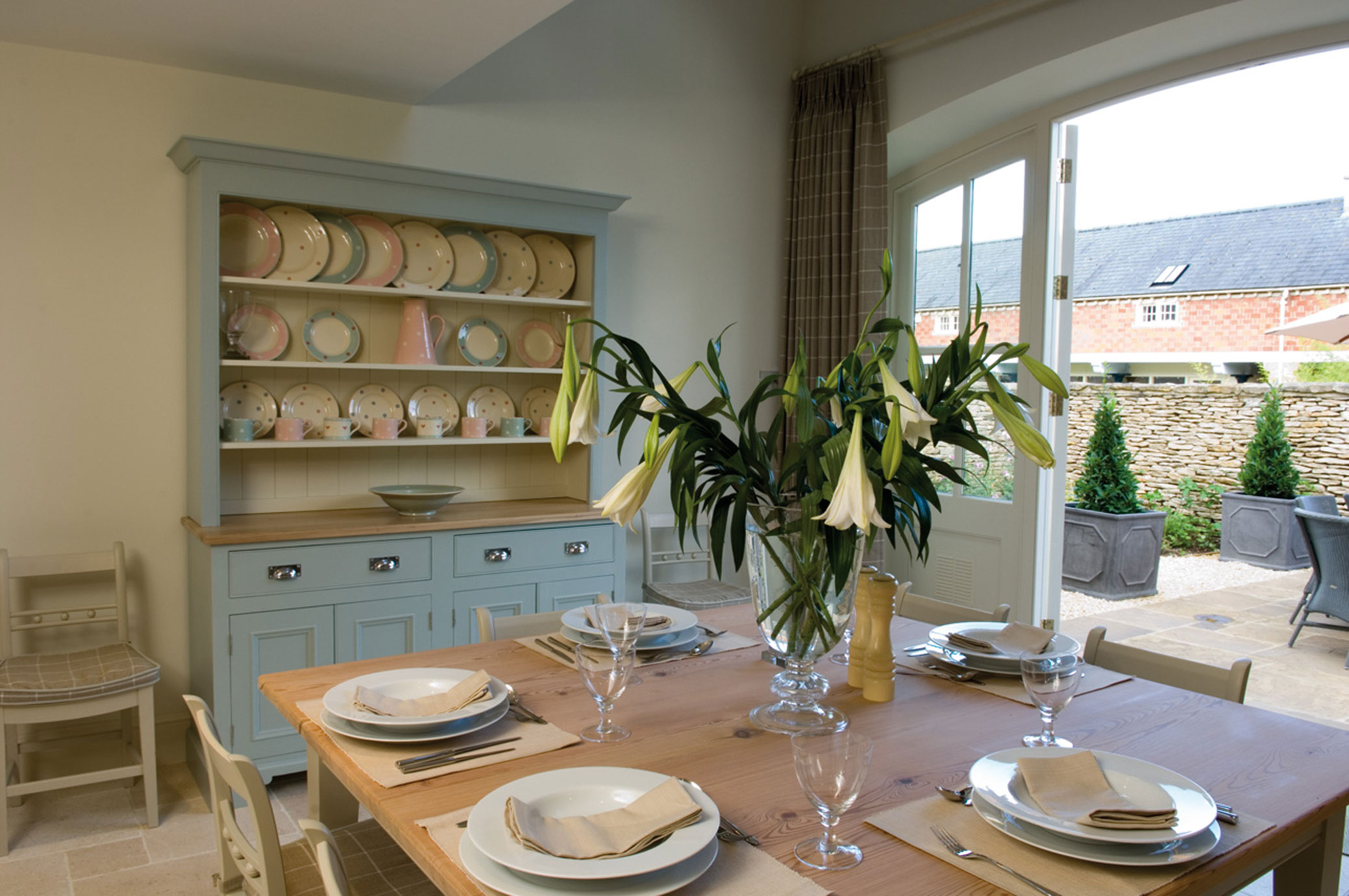 Helles Esszimmer mit Holztisch, vier Gedecken, Vase mit weißen Lilien und Anrichte mit pastellfarbenem Geschirr, offene Tür zum Garten