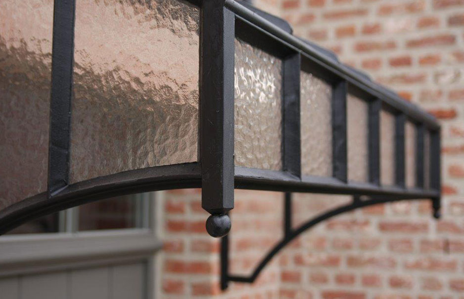 Detailaufnahme eines robusten Vordachs aus schwarzem Schmiedeeisen mit strukturiertem Ornamentglas vor einer Klinkerwand