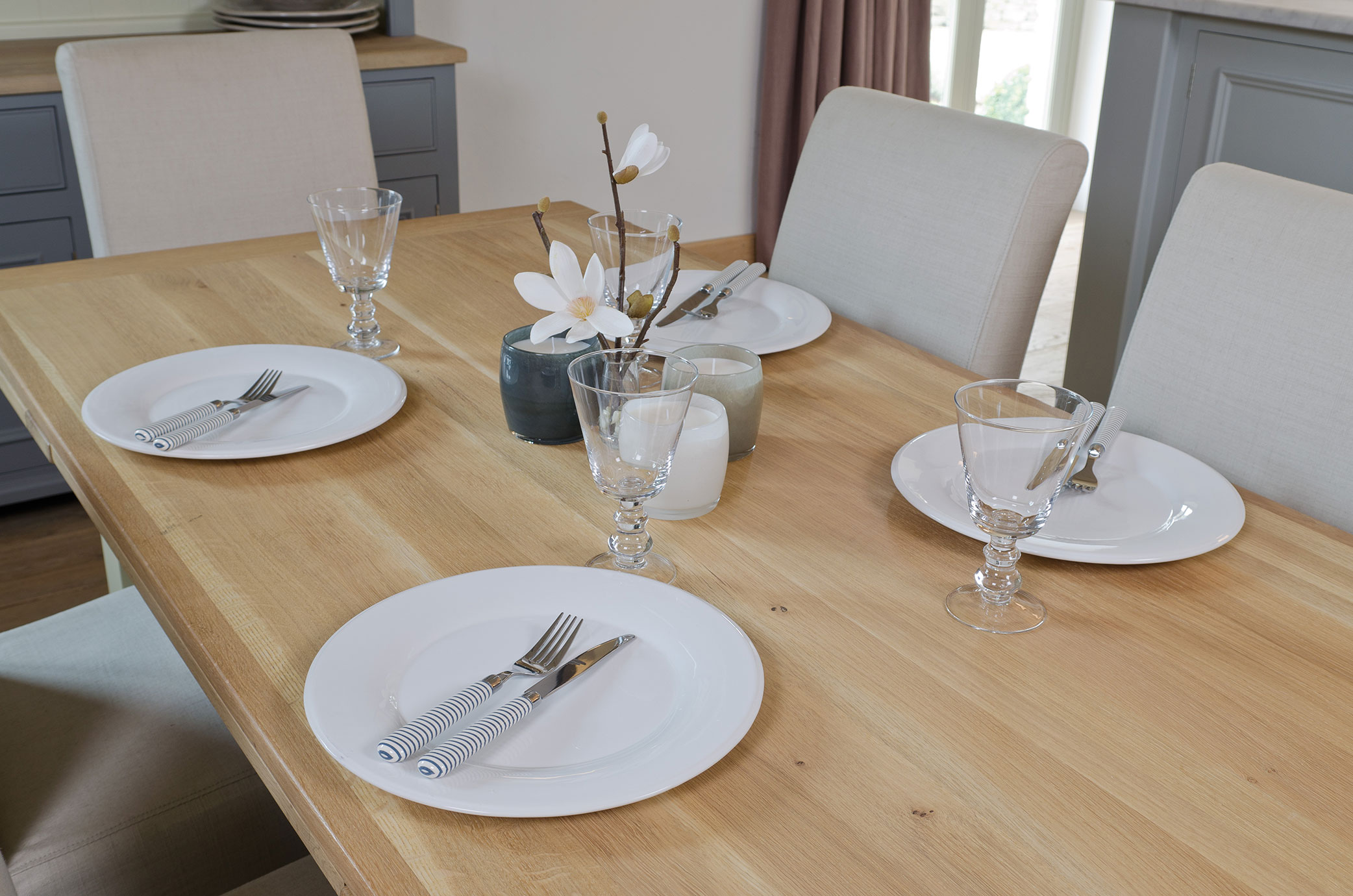 Gedeckter Holzesstisch mit vier weißen Tellern, gestreiftem Besteck, Wassergläsern und minimalistischer Blumendekoration