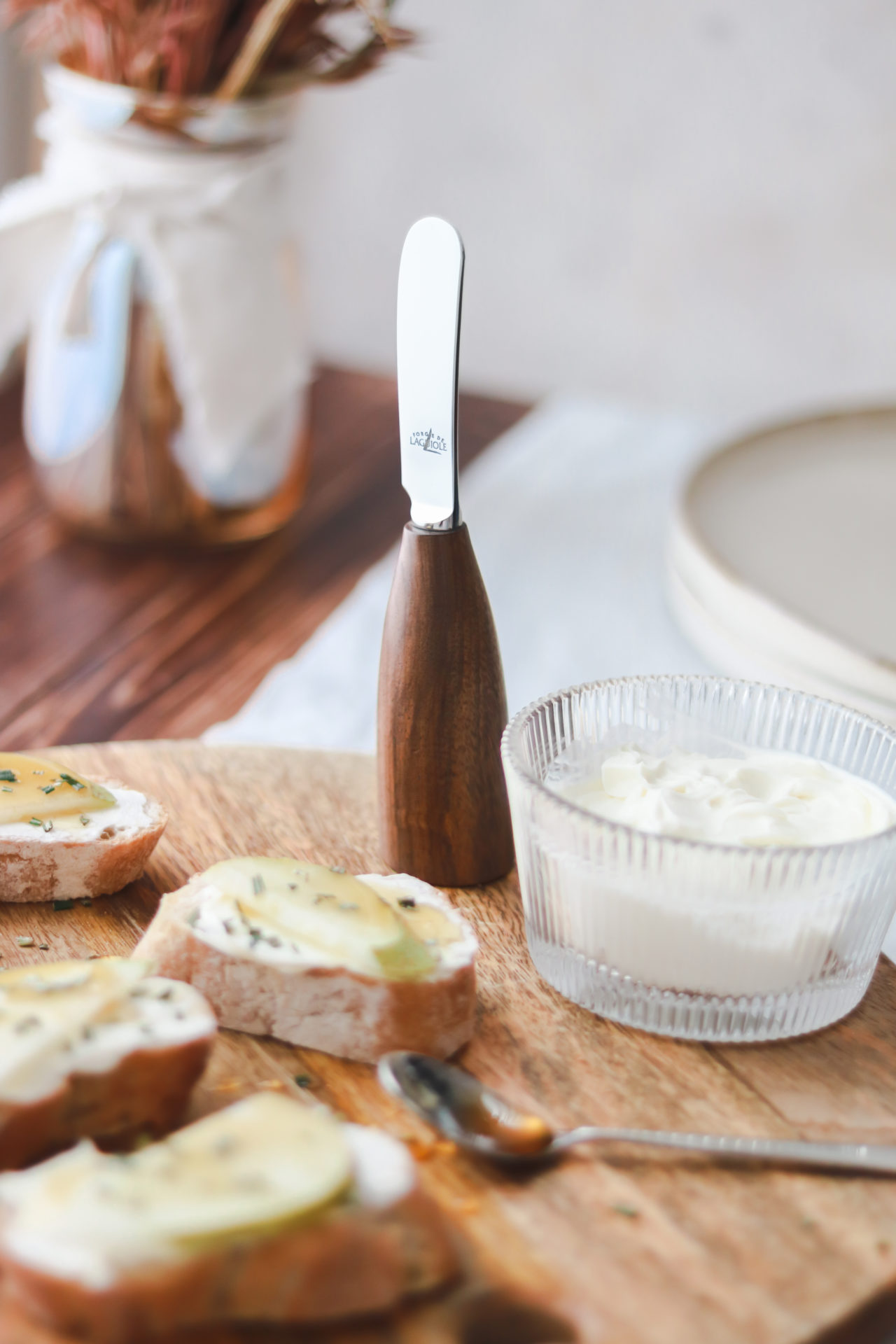 Käsemesser mit dunklem Holzgriff und Edelstahlklinge neben einer Glasschale mit Frischkäse und belegten Brotscheiben auf einem Holzbrett