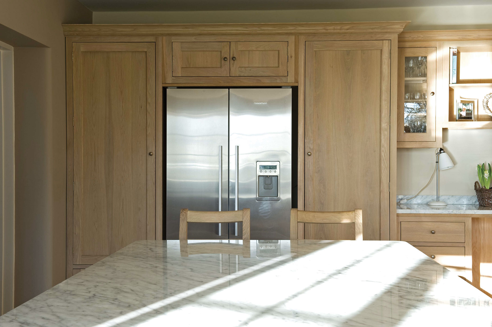 Kühlschrank aus Edelstahl in eine Holzküche integriert, davor Tisch mit Marmorplatte und Holzstühlen