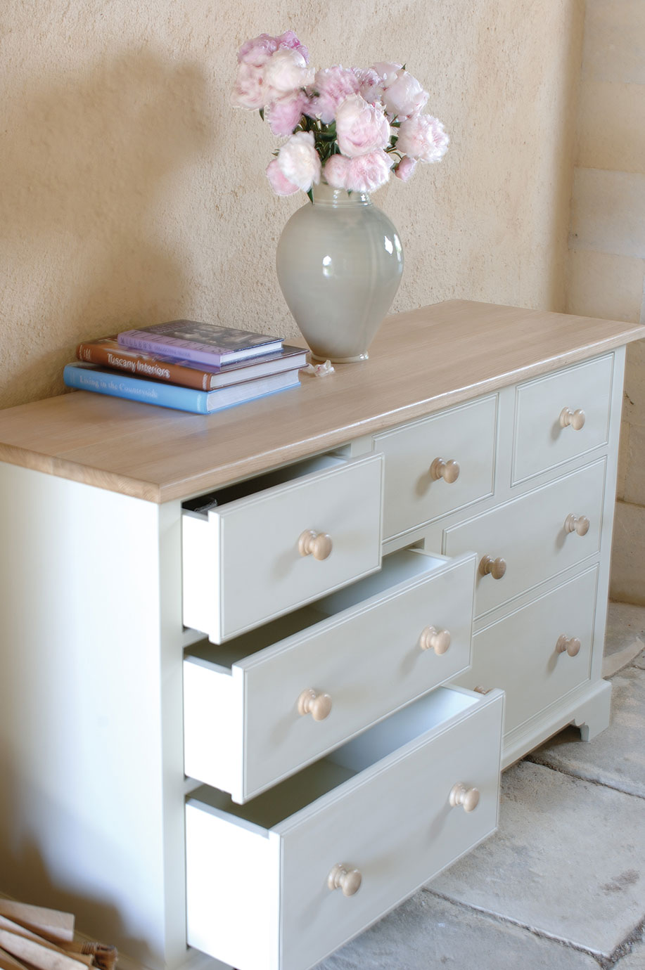 Weiße Kommode mit mehreren offenen Schubladen, darauf eine Vase mit rosa Blumen und ein Stapel Bücher, vor einer beigen Wand stehend