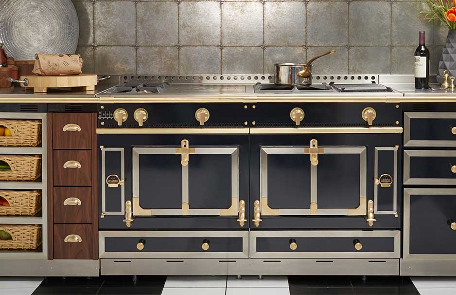 Eleganter Küchenherd im Landhausstil mit dunklem Korpus, goldenen Details, mehreren Backofentüren und Kochfeldern, umgeben von Holzschubladen und Körben