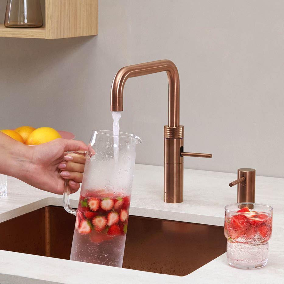 Hand hält eine Glaskaraffe mit Erdbeeren unter einen Kupfer-Wasserhahn in einer modernen Küche