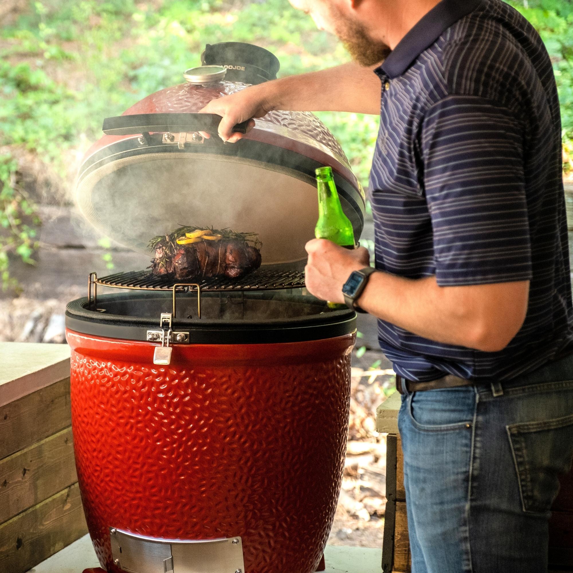 Ein Mann hebt den Deckel eines roten Keramikgrills, während er Fleisch grillt und eine Getränkeflasche hält