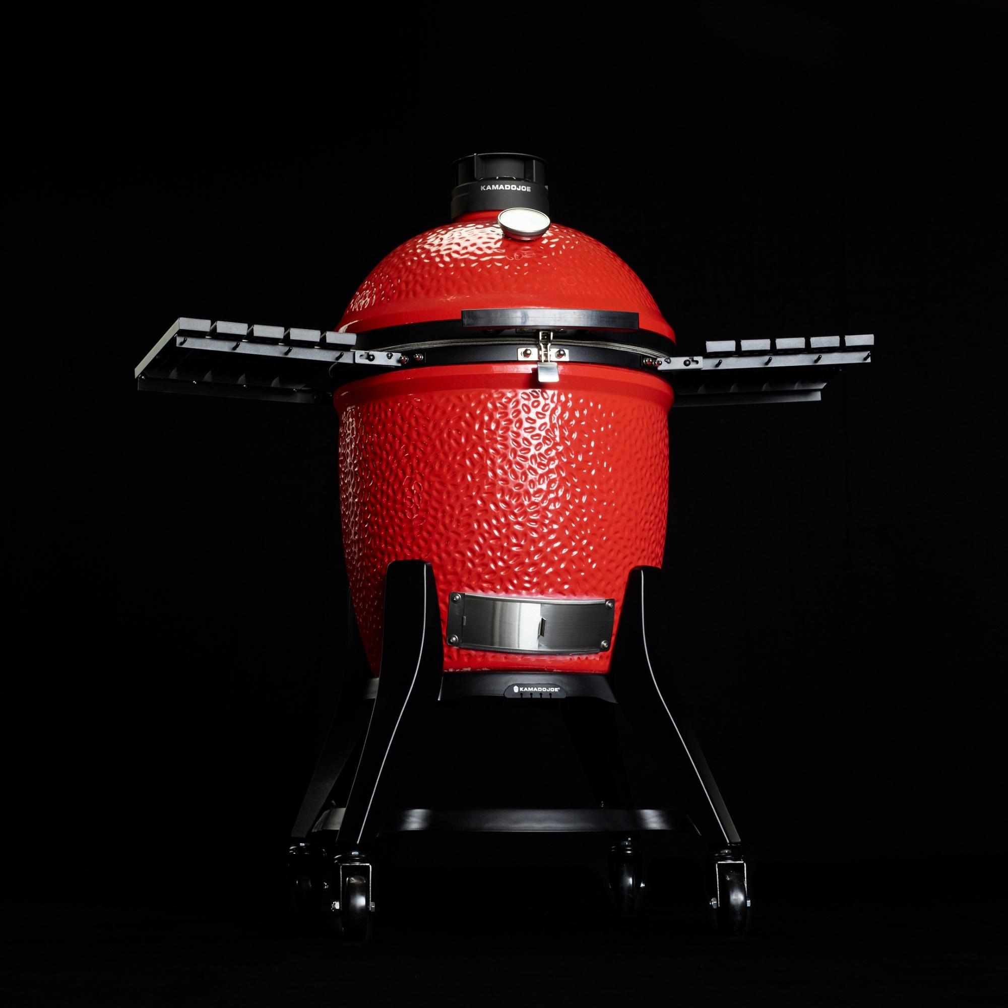 Roter Keramikgrill mit Deckel, seitlichen Ablagen und Standfuß auf Rollen, freigestellt vor schwarzem Hintergrund