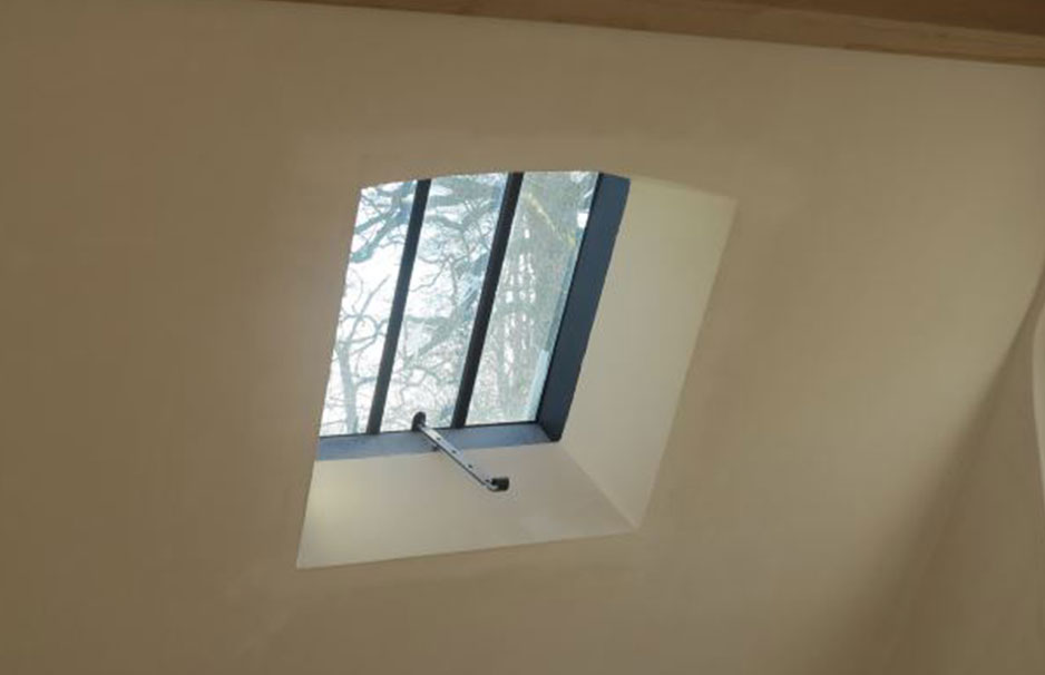 Schräges Dachfenster mit schwarzem Metallrahmen und Griff zum Öffnen, Tageslicht fällt in den Raum, Bäume sind draußen sichtbar.