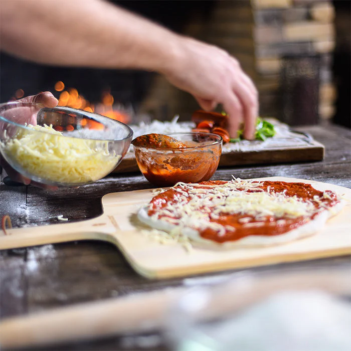 Hand belegt frischen Pizzateig mit Tomatensauce und geriebenem Käse auf einem Holzbrett, Zutaten im Hintergrund