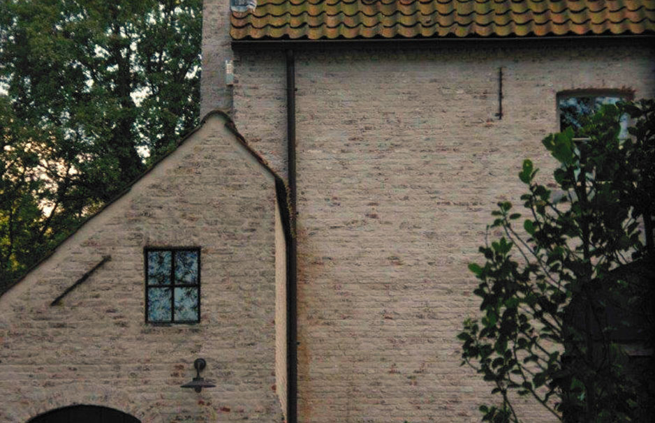 Detailansicht einer Backsteinfassade mit Satteldach, kleinem Fenster und Außenleuchte, teils von Baum verdeckt
