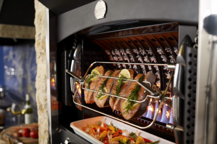 Frische Fischfilets mit Zitronen und Rosmarin werden im hochwertigen Grillofen gegrillt, darunter befindet sich buntes Ofengemüse in einer Form