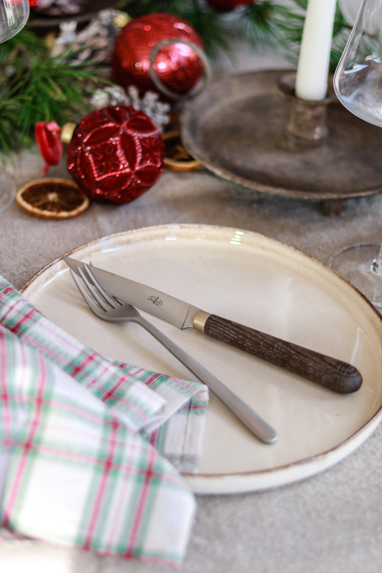 Ein gedeckter Weihnachtstisch mit gezacktem Messer und Gabel auf einem Teller, karierter Serviette, Kerze und roten Weihnachtskugeln als Dekoration