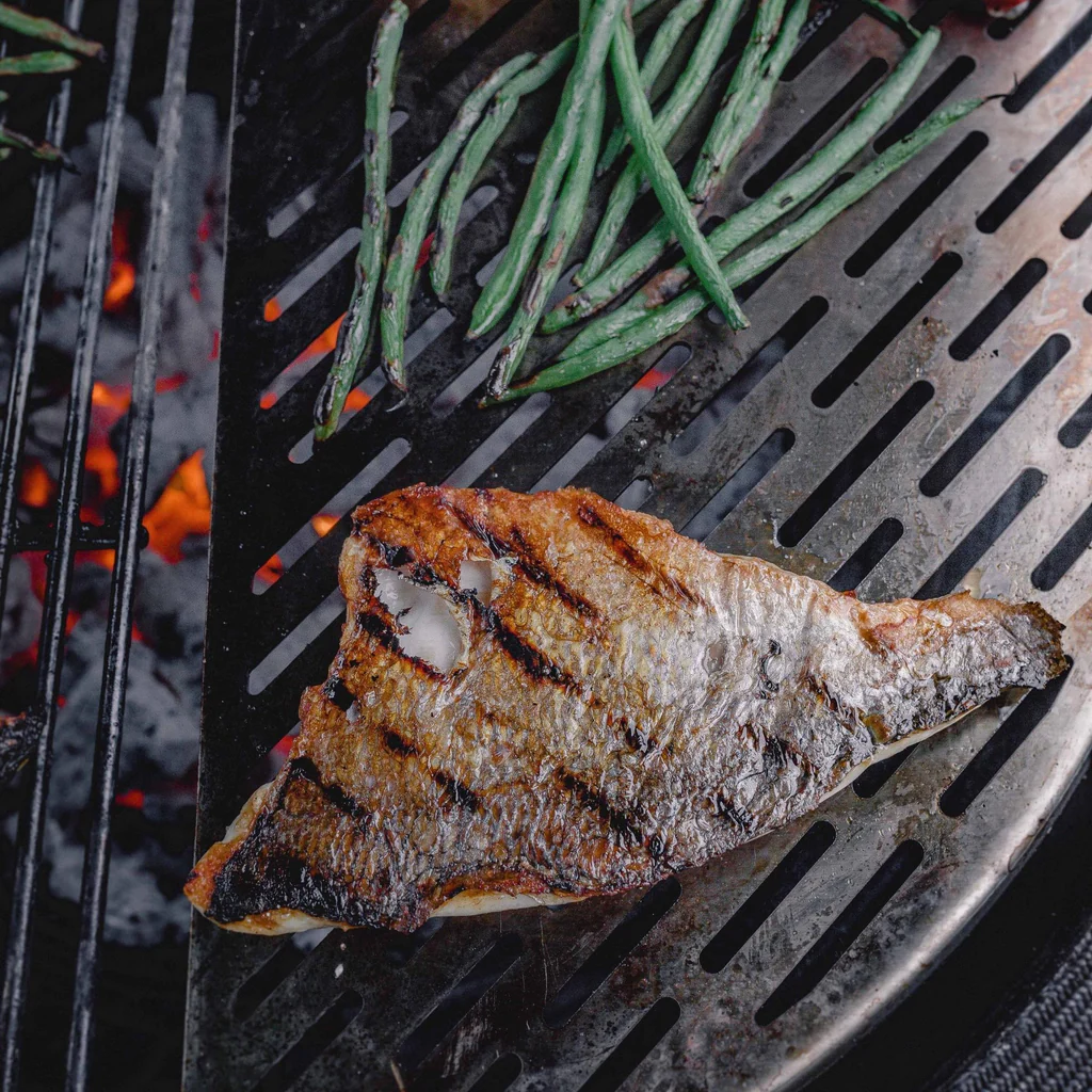 Ein gegrilltes Fischfilet mit knuspriger Haut liegt neben frischen grünen Bohnen auf einem Grill über glühender Holzkohle.