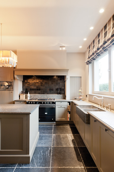 Helle Küche im Landhausstil mit grauen Fronten, Steinboden, großem Fenster und moderner Ausstattung wie Backofen und Spülbecken.
