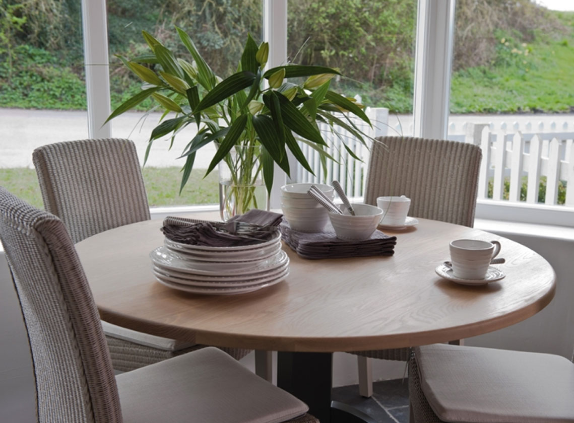 Heller Essbereich mit rundem Holztisch, Gedeck aus Tellern und Schalen, Tassen, Stoffservietten und frischem Blumenstrauß, umgeben von geflochtenen Stühlen