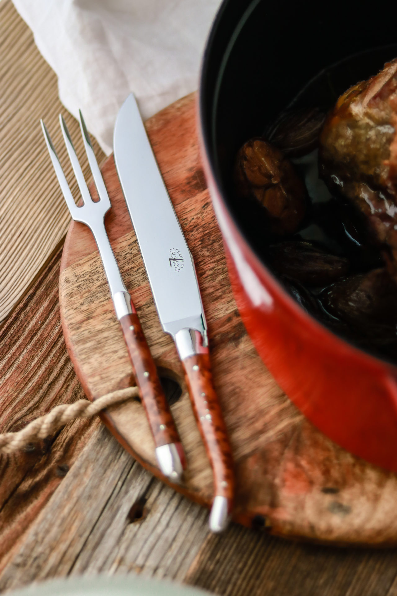 Hochwertiges Tranchiermesser und Fleischgabel mit Holzgriff liegen auf einem runden Schneidebrett neben einem roten Bräter mit Fleisch