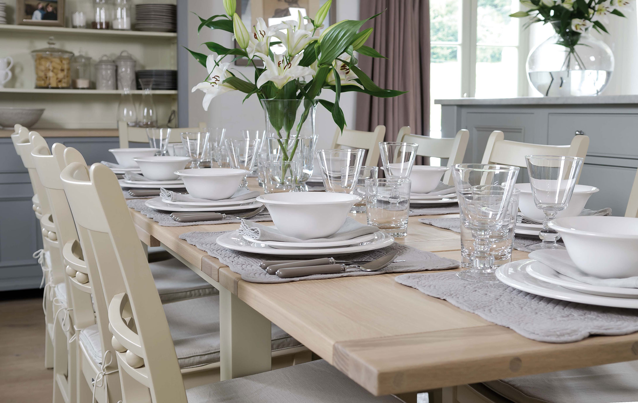 Heller Esstisch im Landhausstil, sorgfältig gedeckt mit weißem Porzellan, Gläsern und Besteck, dazu eine Blumenvase mit Lilien als Mittelpunkt