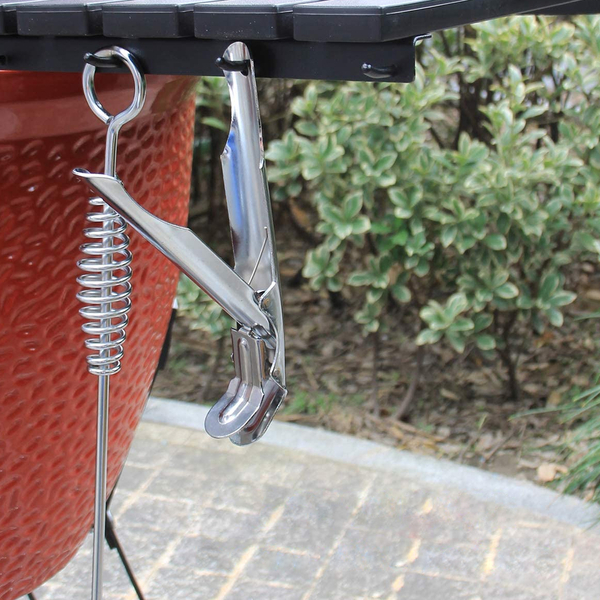 Detailaufnahme einer Edelstahl-Grillzange und einem Grillrostheber, die an einer Seitenablage eines roten Kamado Keramikgrills hängen.