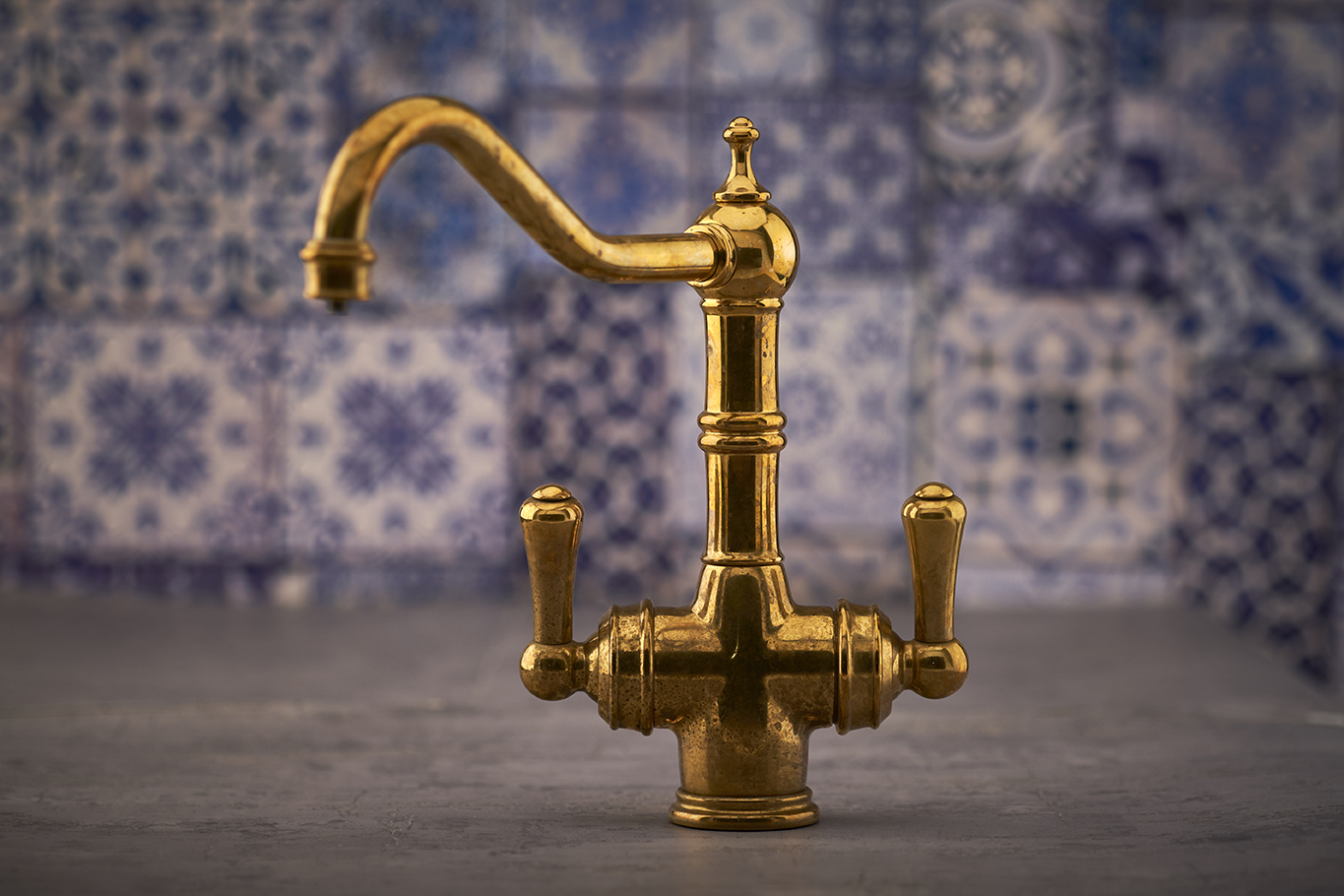 Goldfarbener Vintage-Messing-Wasserhahn mit zwei Griffen und gebogenem Auslauf vor gemusterter Kachelwand