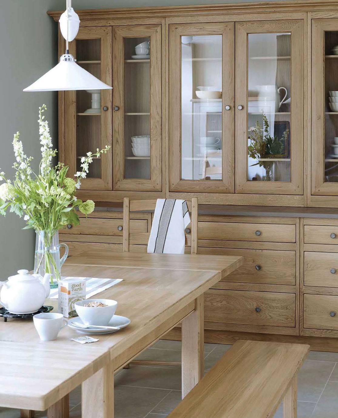 Helles Esszimmer mit Holztisch, Bank, Stuhl, blühenden Blumen und großer Vitrine mit Geschirr