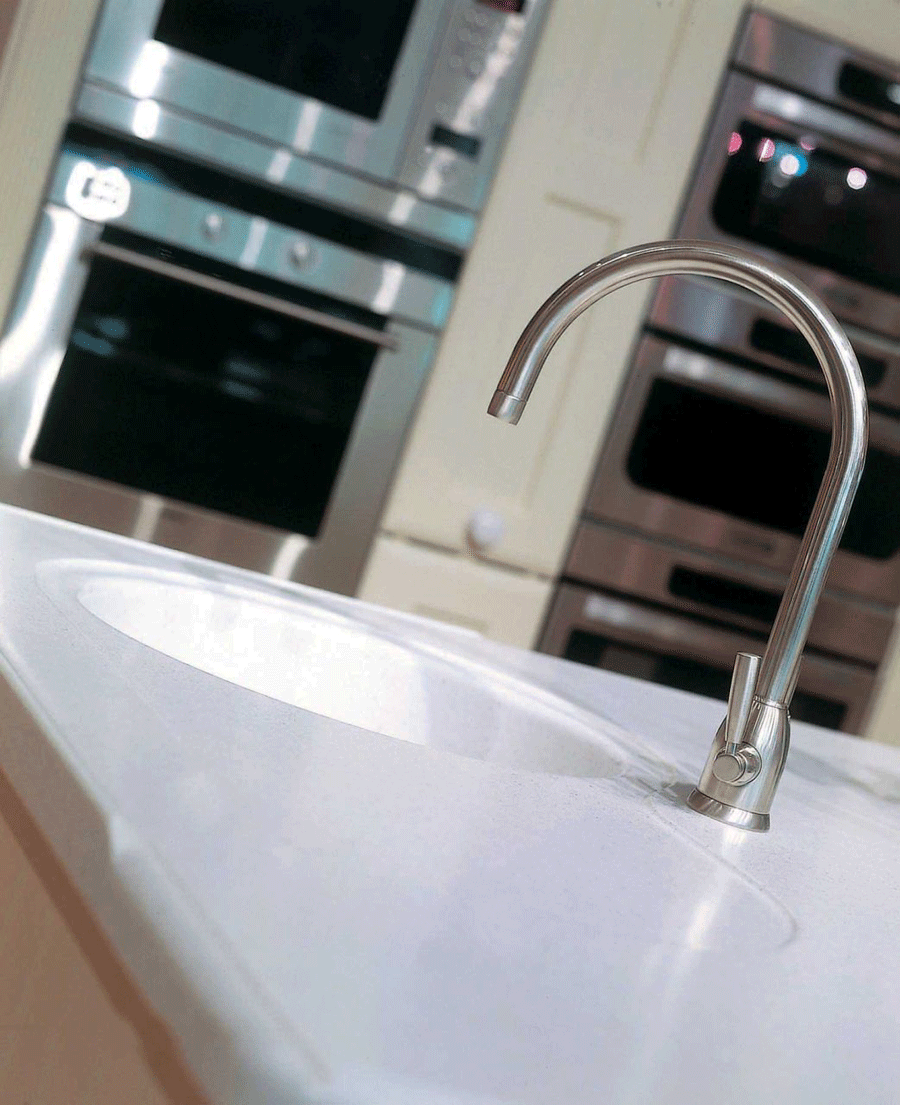 Nahaufnahme einer modernen Küchenarmatur aus Edelstahl über einer weißen Spüle, im Hintergrund hochwertige Einbaugeräte wie Backofen und Mikrowelle.