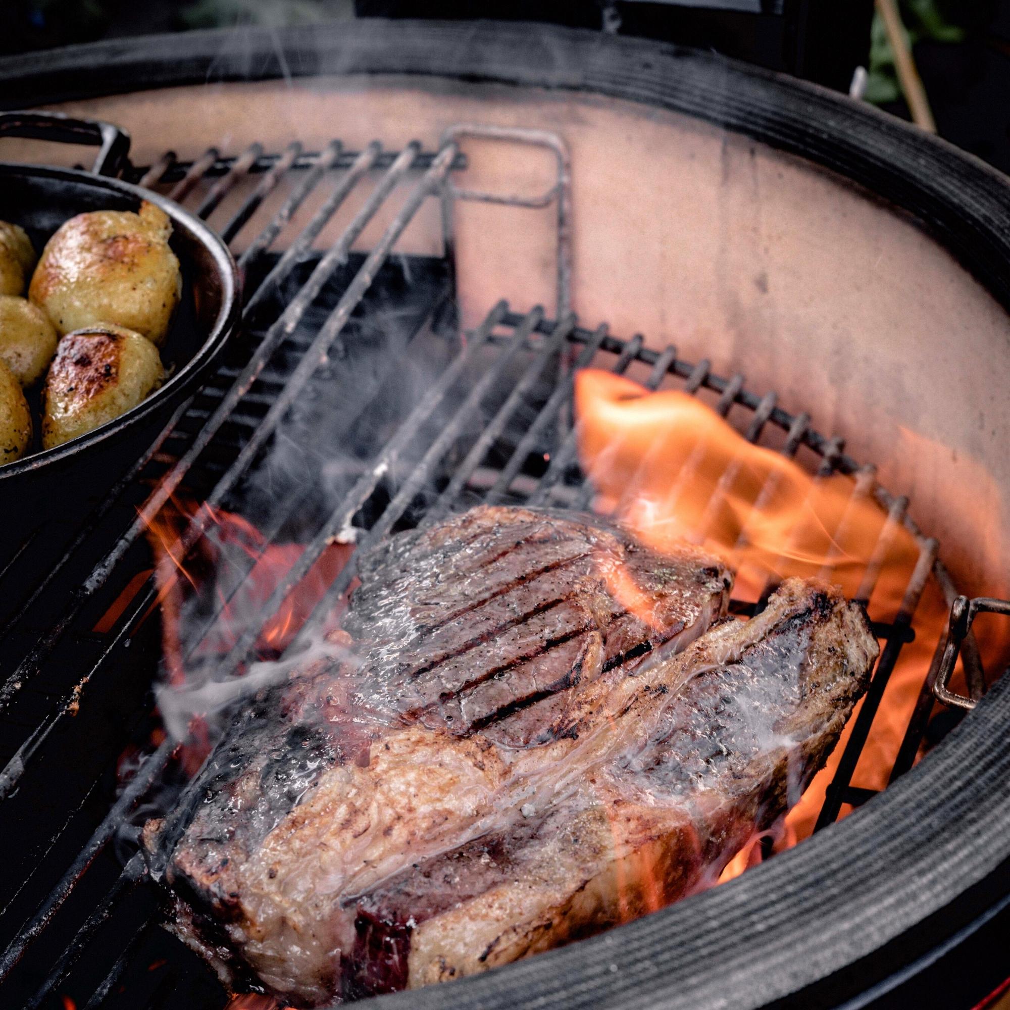 Großes, gegrilltes Steak und Kartoffeln in einer Pfanne auf einem Kugelgrill mit offener Flamme und Rauch