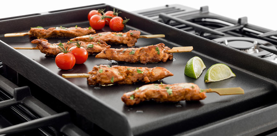 Schwarze Grillplatte auf Gasherd mit gegrillten Hähnchenspießen, frischen Kirschtomaten und Limettenspalten