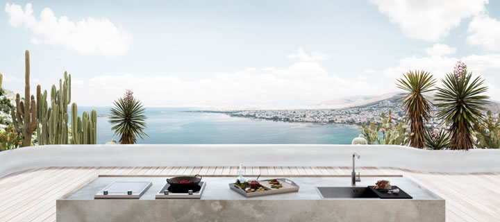 Moderne Außenküche mit Kochfeld, Grill und Spüle auf einer hellen Terrasse, umgeben von Palmen und Kakteen, mit Blick auf das Meer und eine Küstenstadt