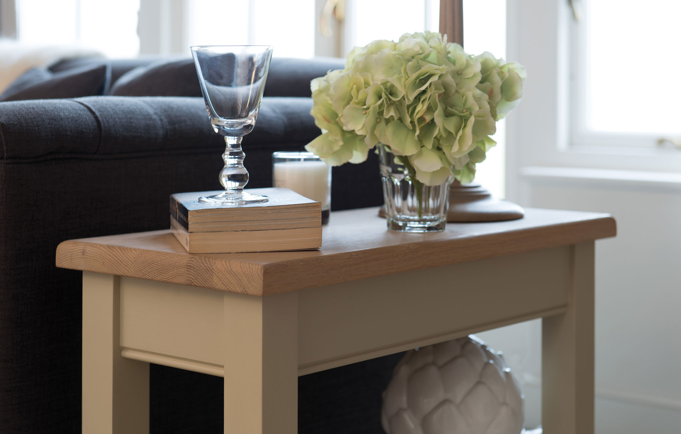 Beistelltisch mit Holzplatte, darauf ein Weinglas, Bücher, eine Kerze und ein Blumenstrauß, daneben ein graues Sofa im Wohnzimmer