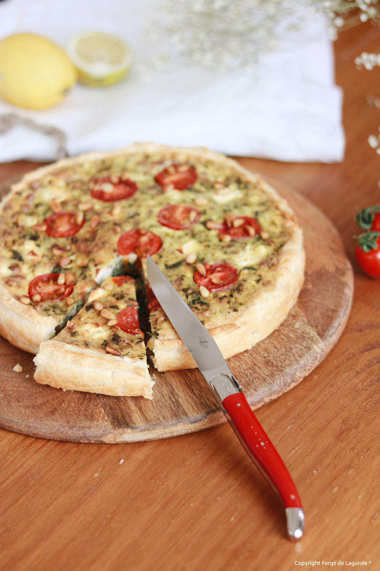 Frisch gebackene Quiche mit Tomaten und Zucchini auf Holzbrett, rotes Laguiole Messer, ein Stück ist geschnitten