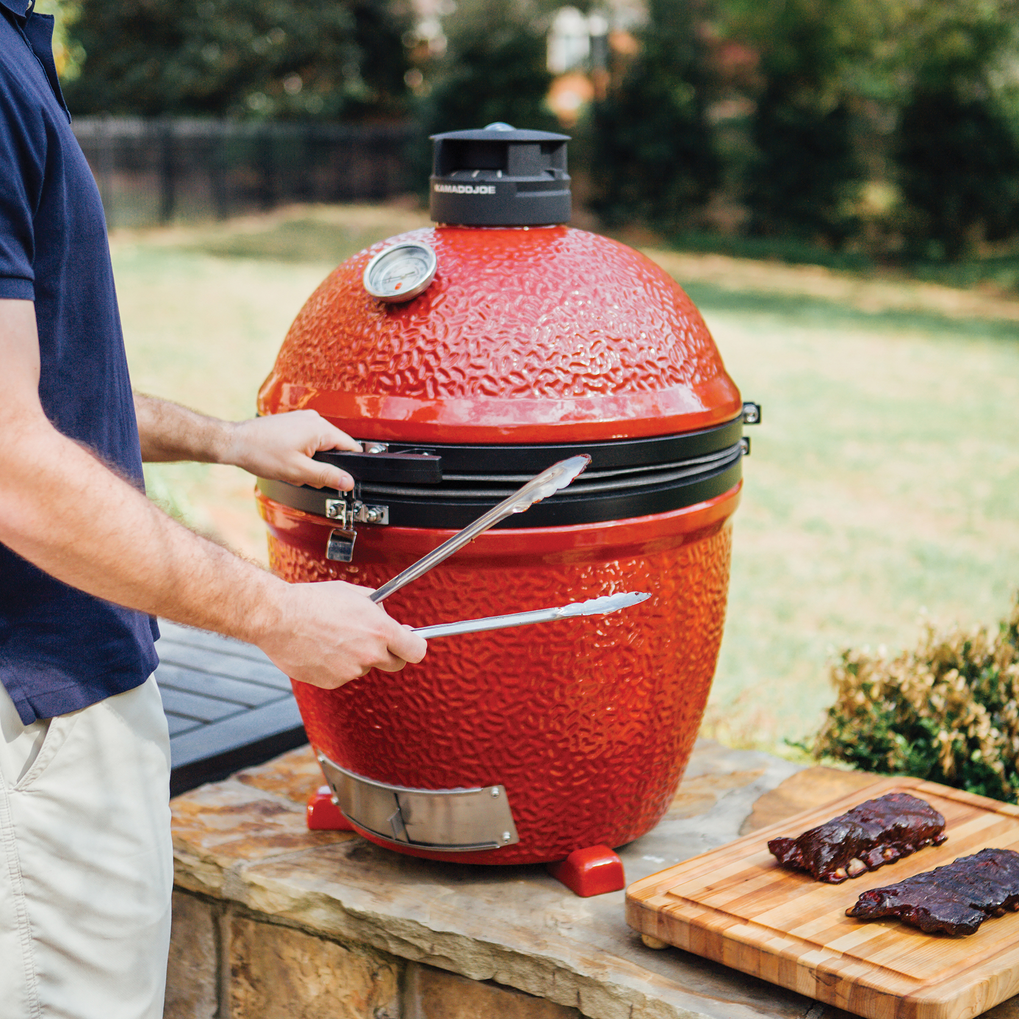 Ein roter Kamado Joe Keramikgrill steht im Garten, daneben hält eine Person Grillzangen. Auf einem Holzbrett liegen zwei Portionen gegrillte Rippchen.