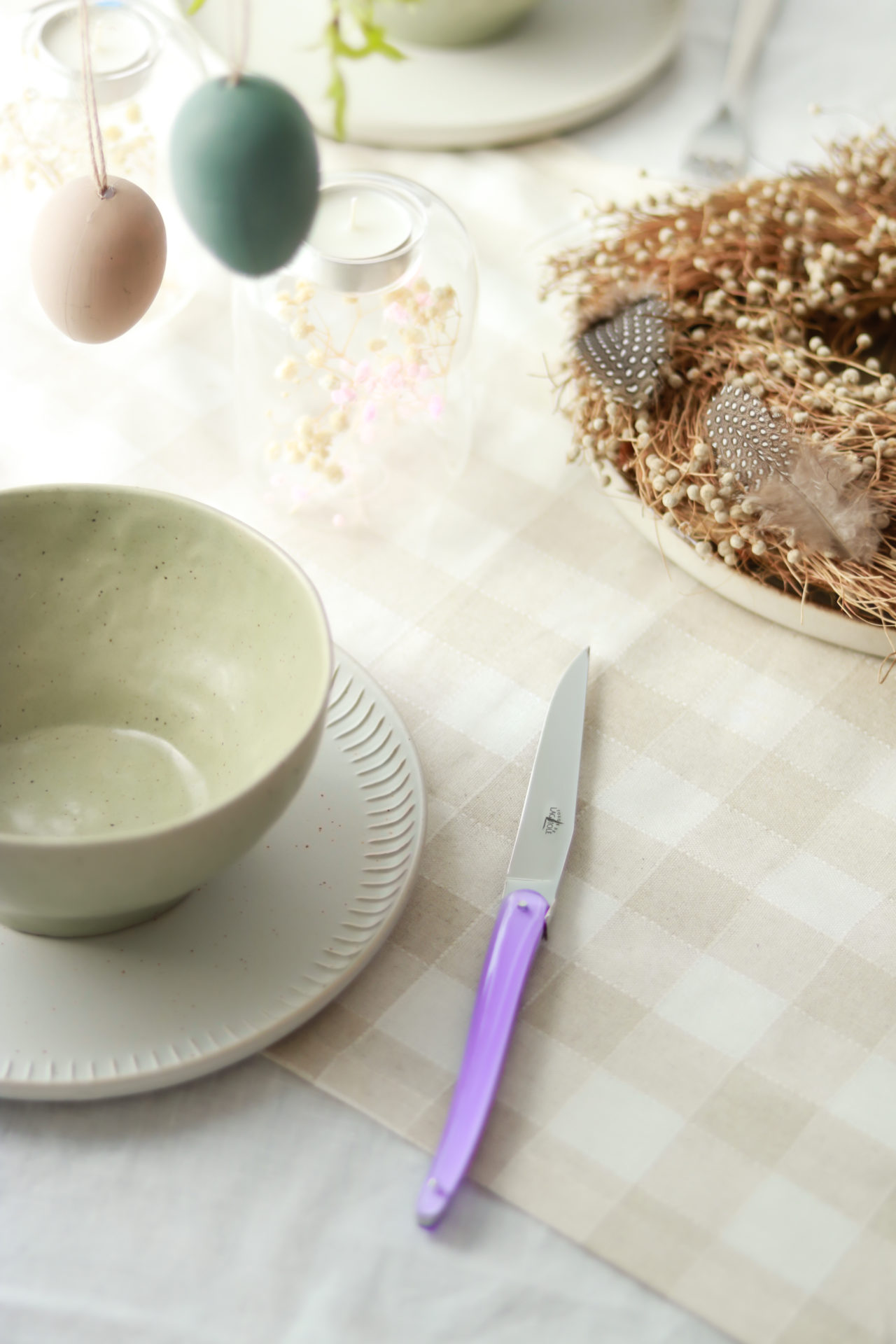 Detail eines österlich dekorierten Tisches mit pastellfarbener Keramikschale, Messer mit lila Griff, Deko-Eiern, Kerze und geflochtenem Nest mit Federn