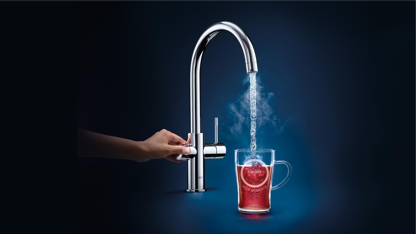Hand bedient einen Grohe Wasserhahn, der kochend heißes Wasser in ein Teeglas gießt, aus dem Dampf aufsteigt