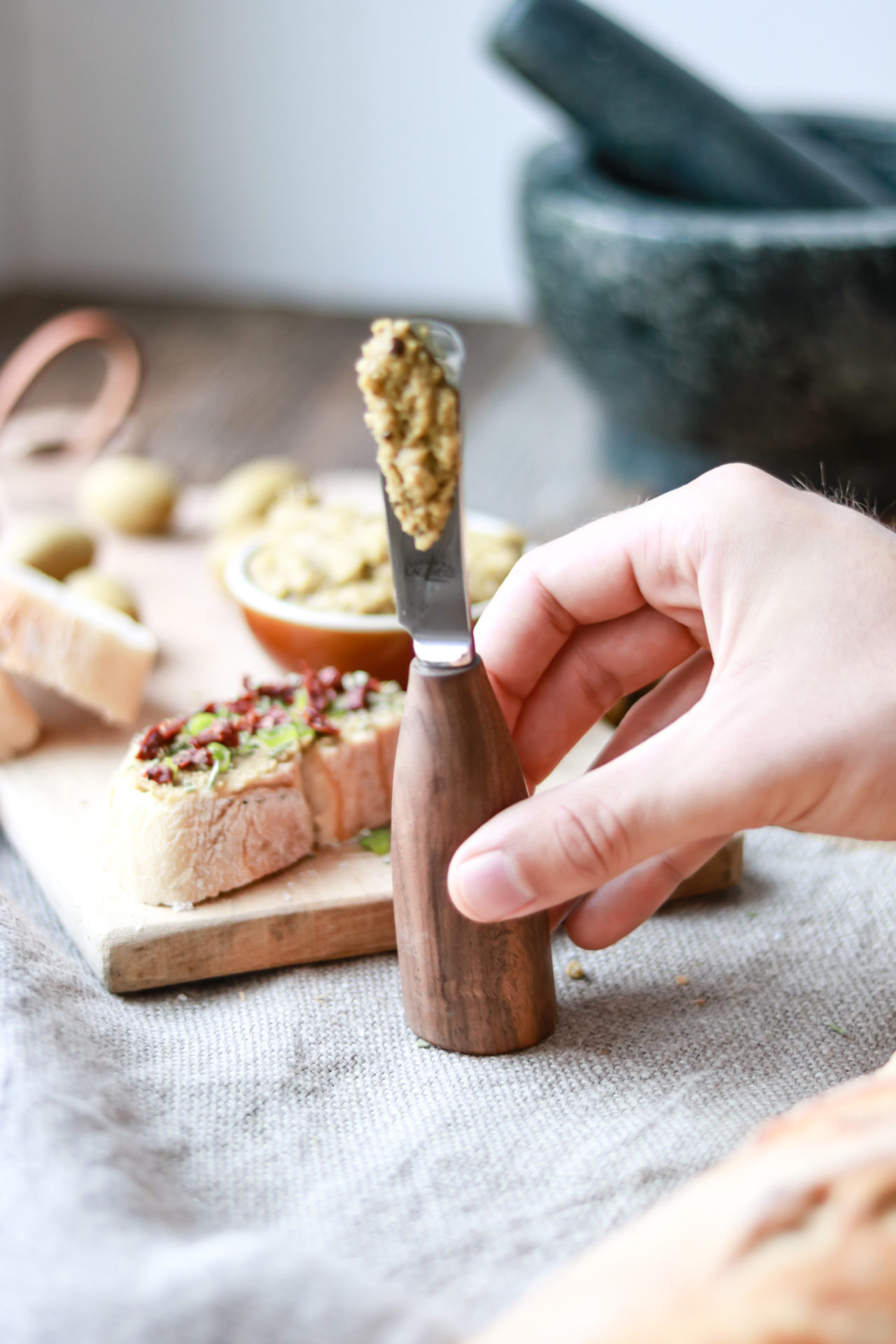 Hand hält ein Messer mit Holzgriff und streicht grüne Tapenade auf Baguette; im Hintergrund Brot und Oliven