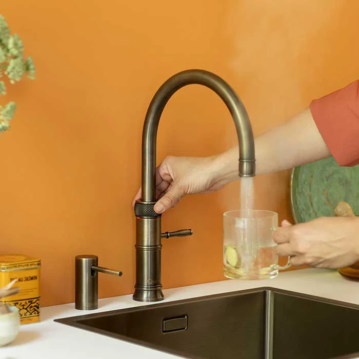 Hand bedient eine bronzefarbene Küchenarmatur und füllt eine Glas-Tasse mit heißem Wasser an einer modernen Spüle vor orangefarbener Wand