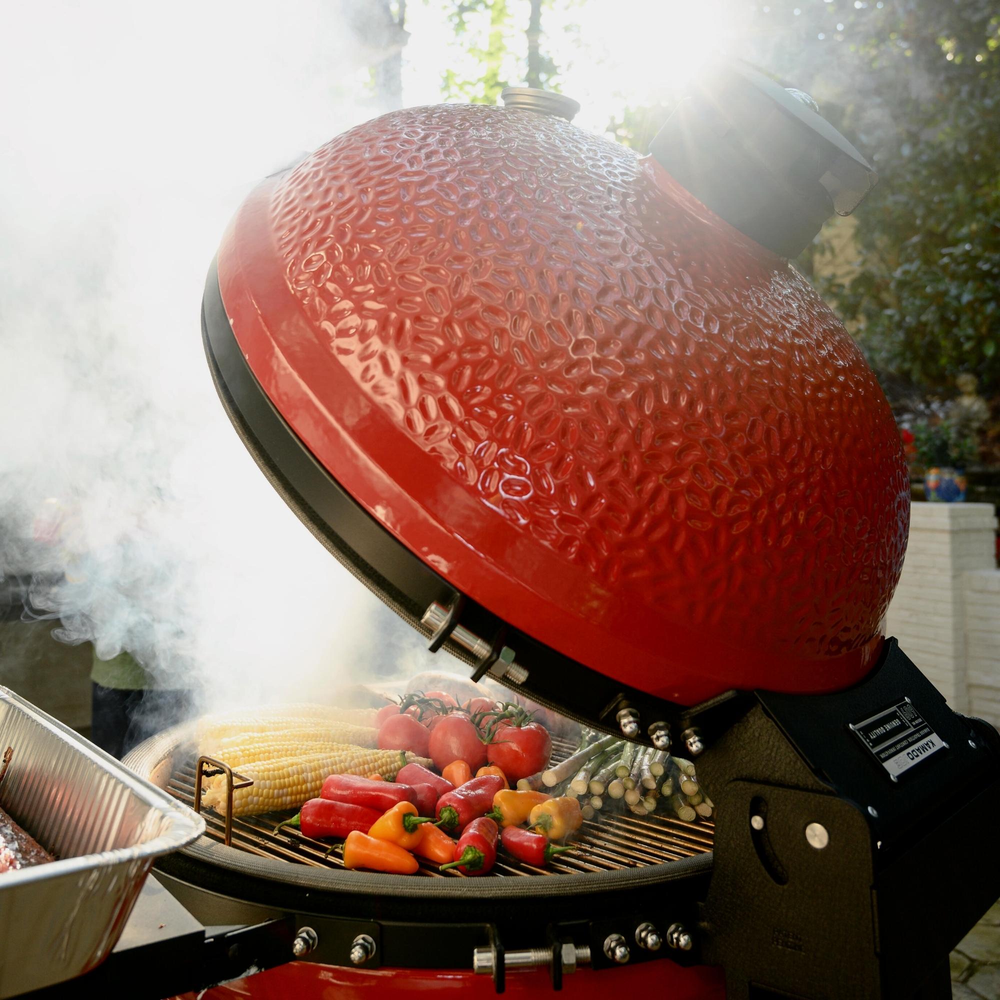 Offener roter Keramikgrill mit verschiedenen Gemüsesorten wie Mais, Paprika, Tomaten und Spargel, der während des Grillens stark raucht