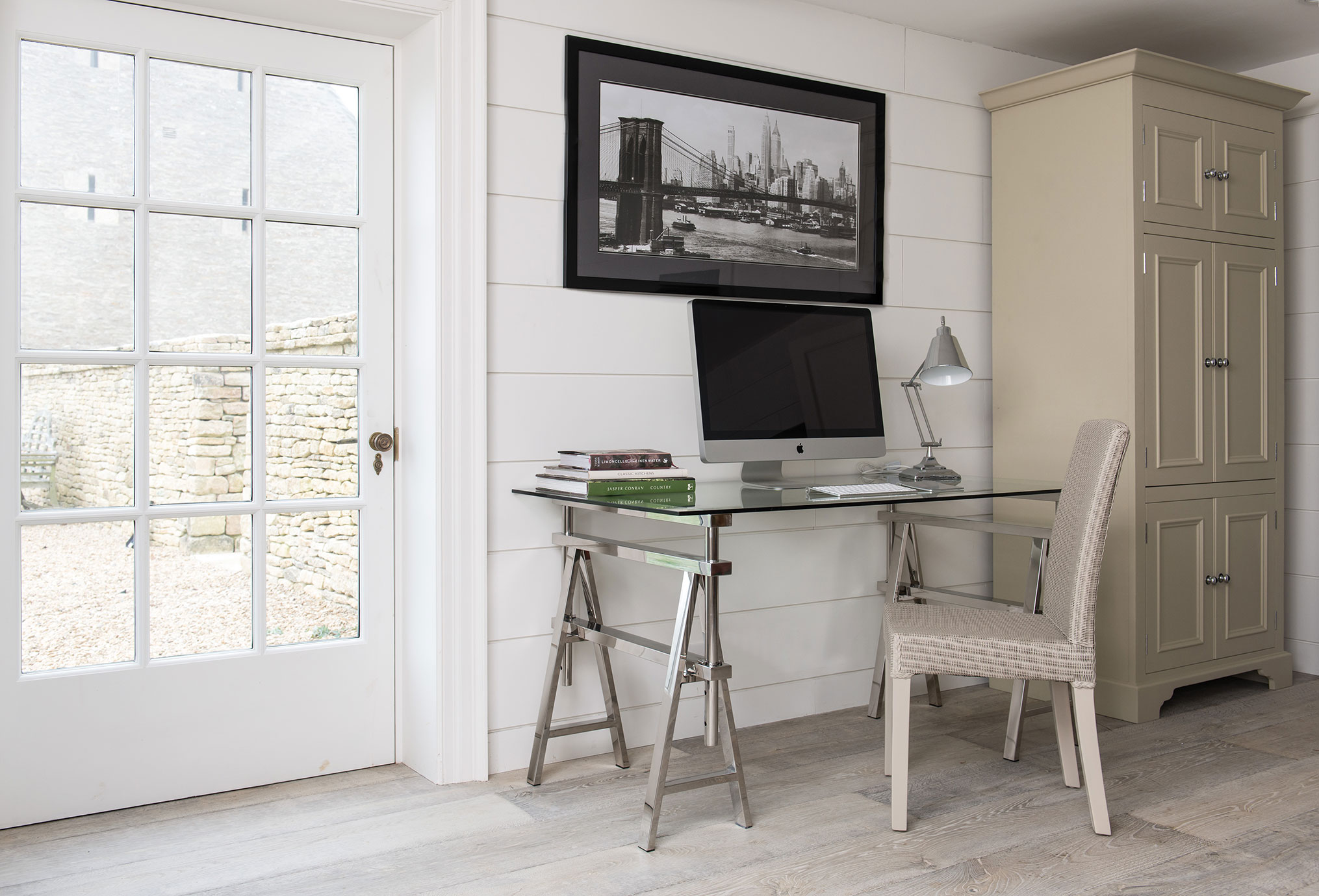 Helles Homeoffice mit Glastisch, Computer, Büchern, Leselampe, gepolstertem Stuhl und Schrank vor weißer Holzwand