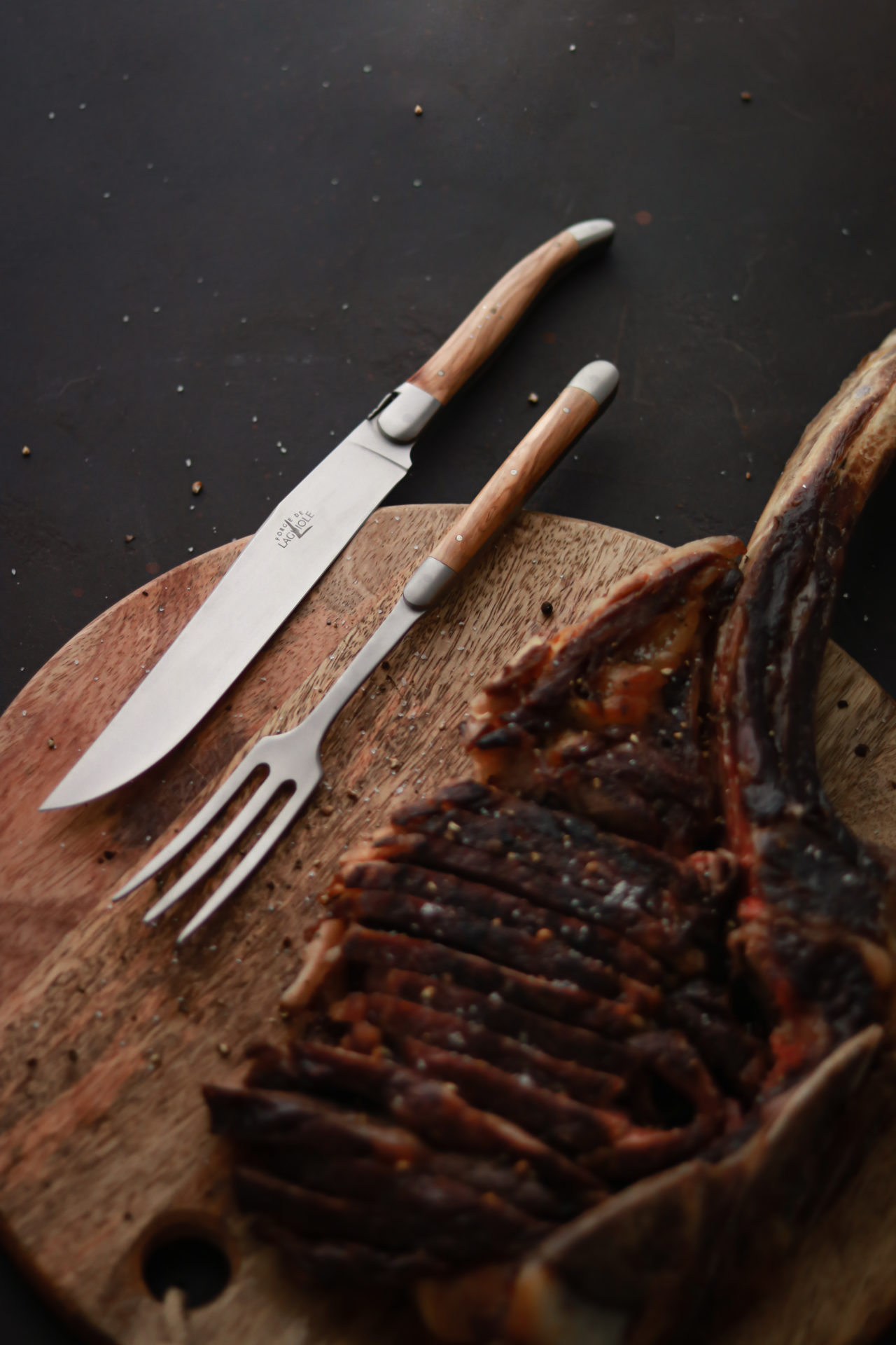 Steakmesser und Fleischgabel mit Holzgriff liegen auf einem runden Holzbrett, neben einem aufgeschnittenen, gewürzten Tomahawk-Steak