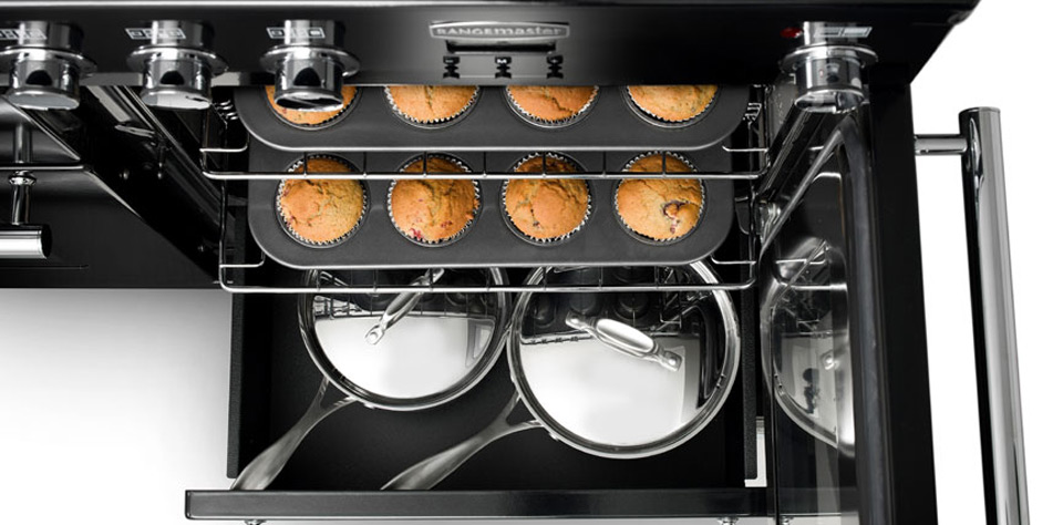 Offener Backofen mit einem Muffinblech, auf dem acht Muffins gebacken werden, darunter befinden sich zwei glänzende Kochtöpfe mit Deckel auf einem Blech.
