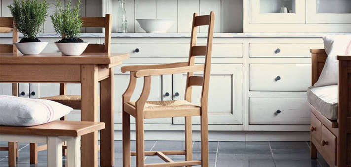Freundliches Esszimmer mit Holztisch, Stühlen, Bank und weißer Landhausküche im Hintergrund. Pflanzen als Dekoration auf dem Tisch.